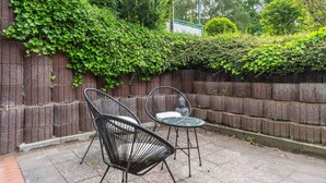 Un patio al aire libre con dos sillas de metal y una mesa redonda pequeña, rodeado por un muro de ladrillo y abundante vegetación.