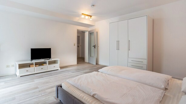 Un dormitorio con una cama, un televisor sobre un mueble bajo, un armario y una puerta que da a otra habitación.