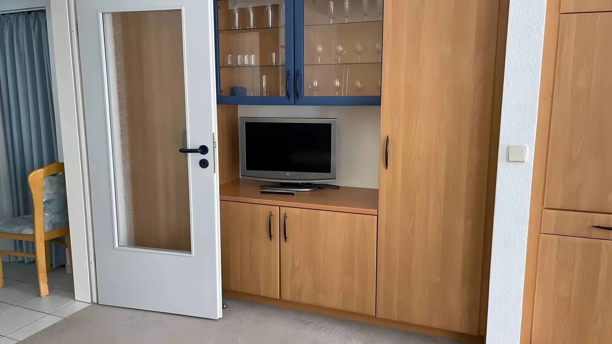 Een kamer met een televisie op een houten kast, een glazen kast met wijnglazen, en een deur met een ruit.
