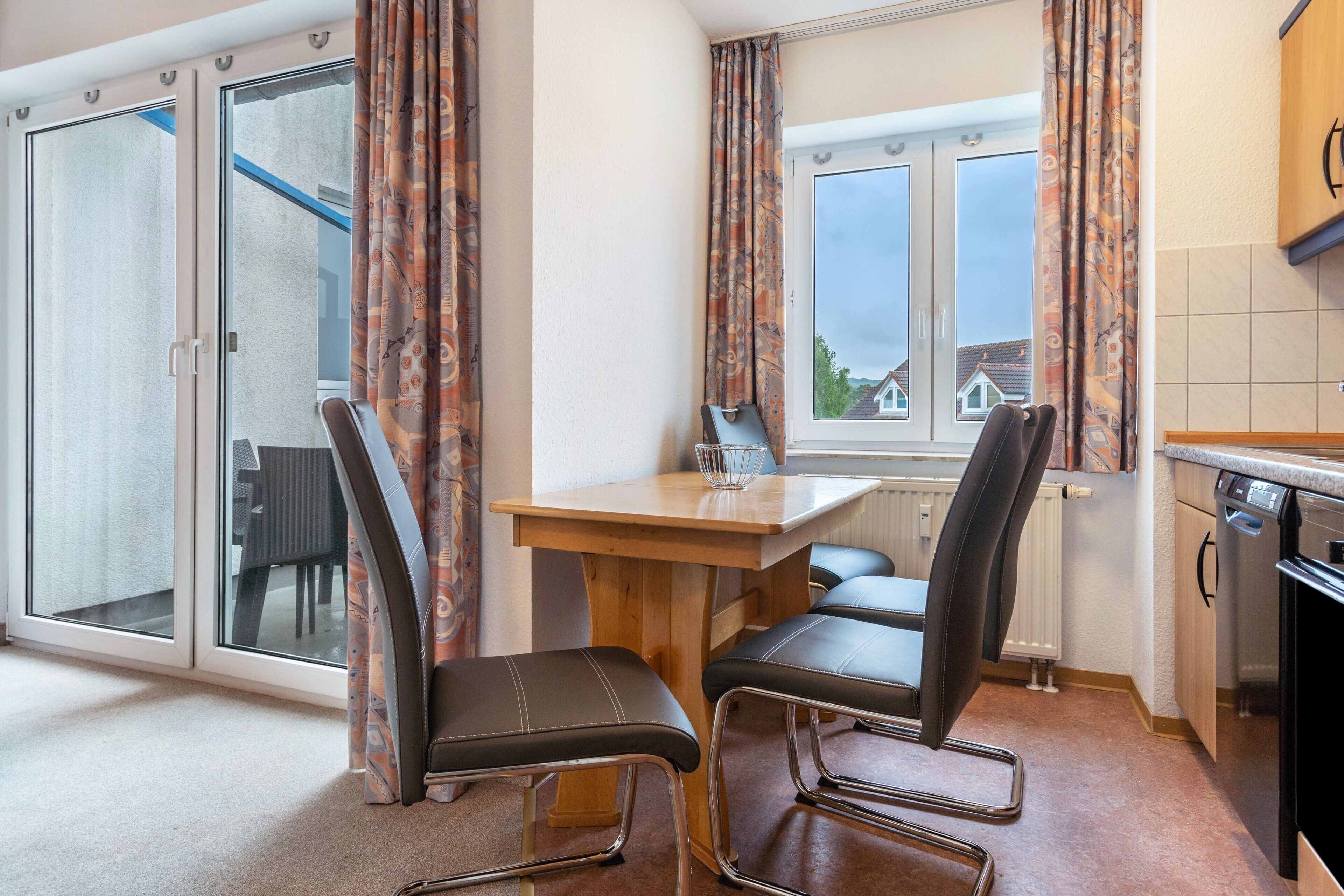 Une cuisine avec une table à manger, des chaises, une fenêtre avec des rideaux et une vue sur les maisons à l’extérieur.