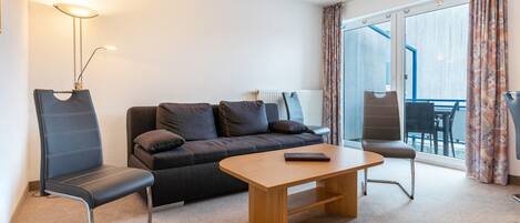 A modern hotel room with a sofa, two chairs, a wooden coffee table, a lamp, and a view through a glass door.