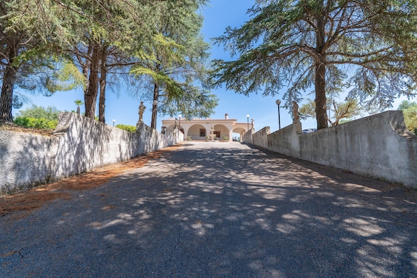 A paved pathway leading to a building with a tiled roof, flanked by trees and stone walls.