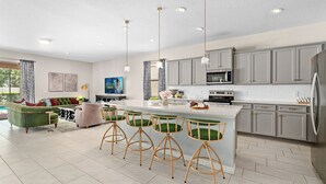 A modern kitchen with grey cabinets, a central island with bar stools, a dining area with a green sofa, and a large window. - NEW! Princess Theme! Minutes to Disney World! Private Pool and Hot tub! Theatre! (Kissimmee)