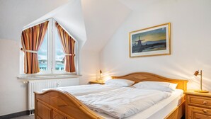 Quarto com cama de madeira, janela com cortinas, mesa de cabeceira com abajur e um quadro na parede.