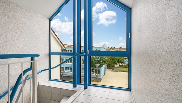 Escada com corrimão azul que leva a uma grande janela com vista clara para prédios lá fora.