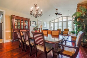 Une salle à manger avec une grande table en bois, six chaises, un lustre et un ventilateur de plafond.