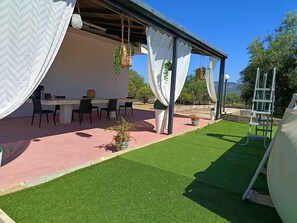 Eine Terrassenanlage im Freien mit Tisch und Stühlen, eine Pergola mit weißen Vorhängen sowie eine Spielgeräte-Anlage.