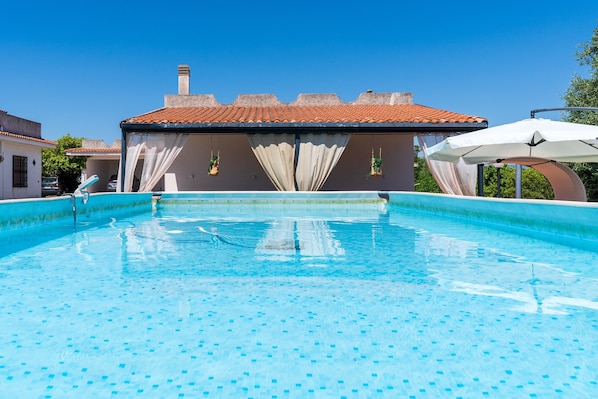 Una piscina con solárium empedrado, un edificio con techo de tejas y un cielo azul despejado.