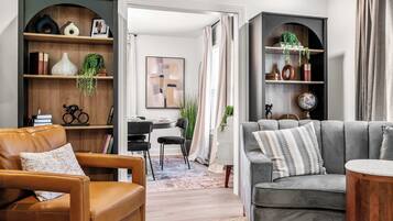 A modern living room with a leather sofa, a grey armchair, and a wooden coffee table. There is a bookshelf with various decorative items and a dining area with a table and chairs.