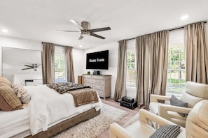 Ein modernes Schlafzimmer mit Bett, Fernseher, Deckenventilator und Blick auf Grünfläche.