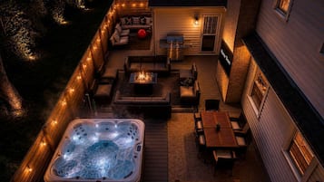 Une terrasse dotée d’un jacuzzi, d’un espace salon et d’un coin barbecue, le tout entouré d’une terrasse en bois et illuminé de guirlandes lumineuses.