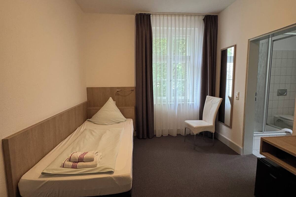 Habitación de hotel con cama, silla, escritorio, espejo y zona de ducha.