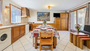 Een keuken met een eettafel voor vier personen, houten kastjes, een wasmachine en een groot raam met gordijnen.