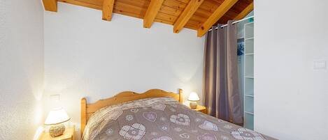 A bedroom with a wooden ceiling, a bed with a patterned bedspread, bedside lamps, and a window with curtains.