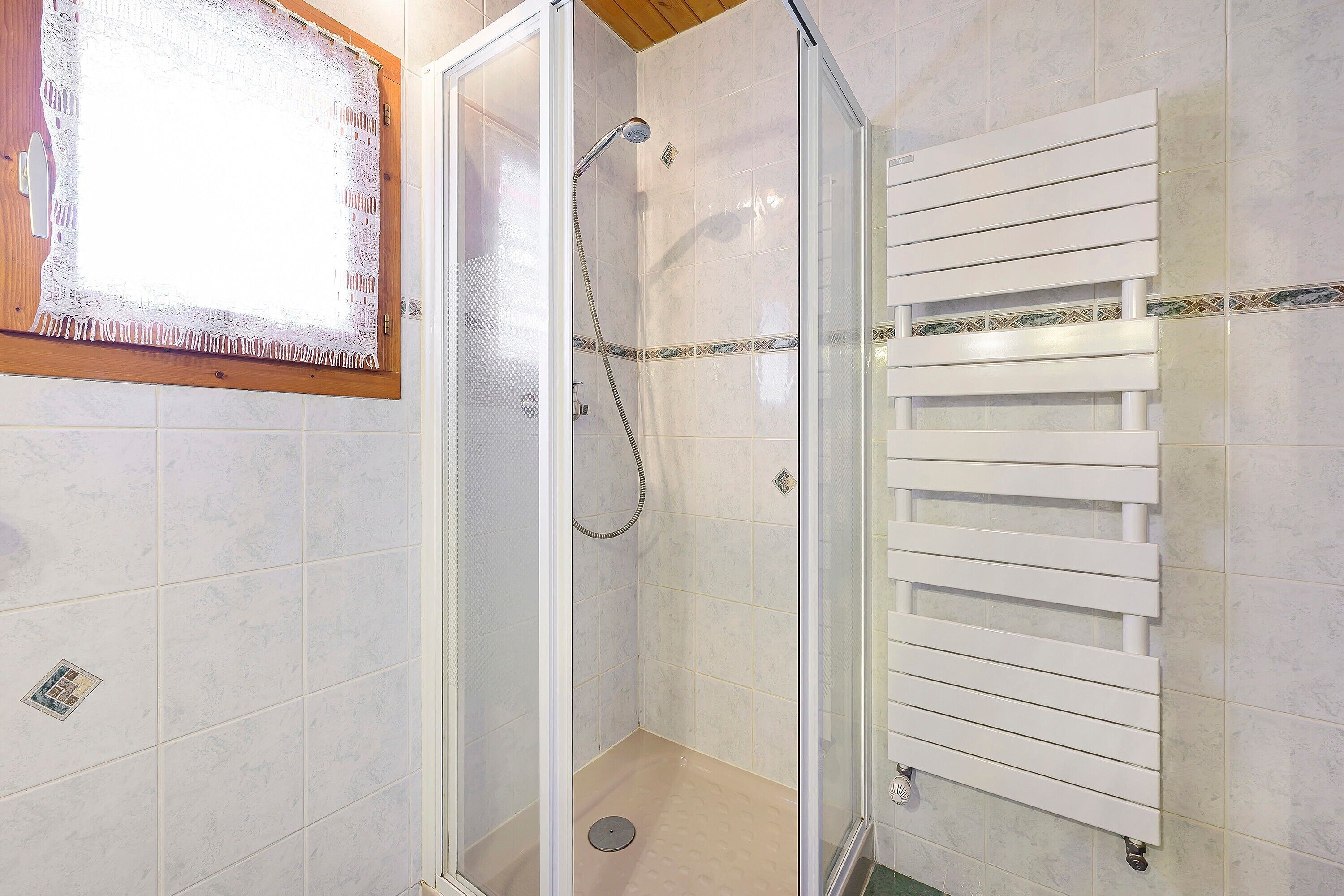 Una ducha con puerta de cristal, un perchero blanco y un ventanal con marco de madera.