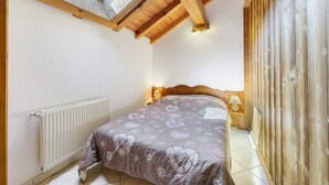 Een slaapkamer met een schuine kap, houten balken, een bed met een beddeken met dessin, nachtlampjes en een radiator.