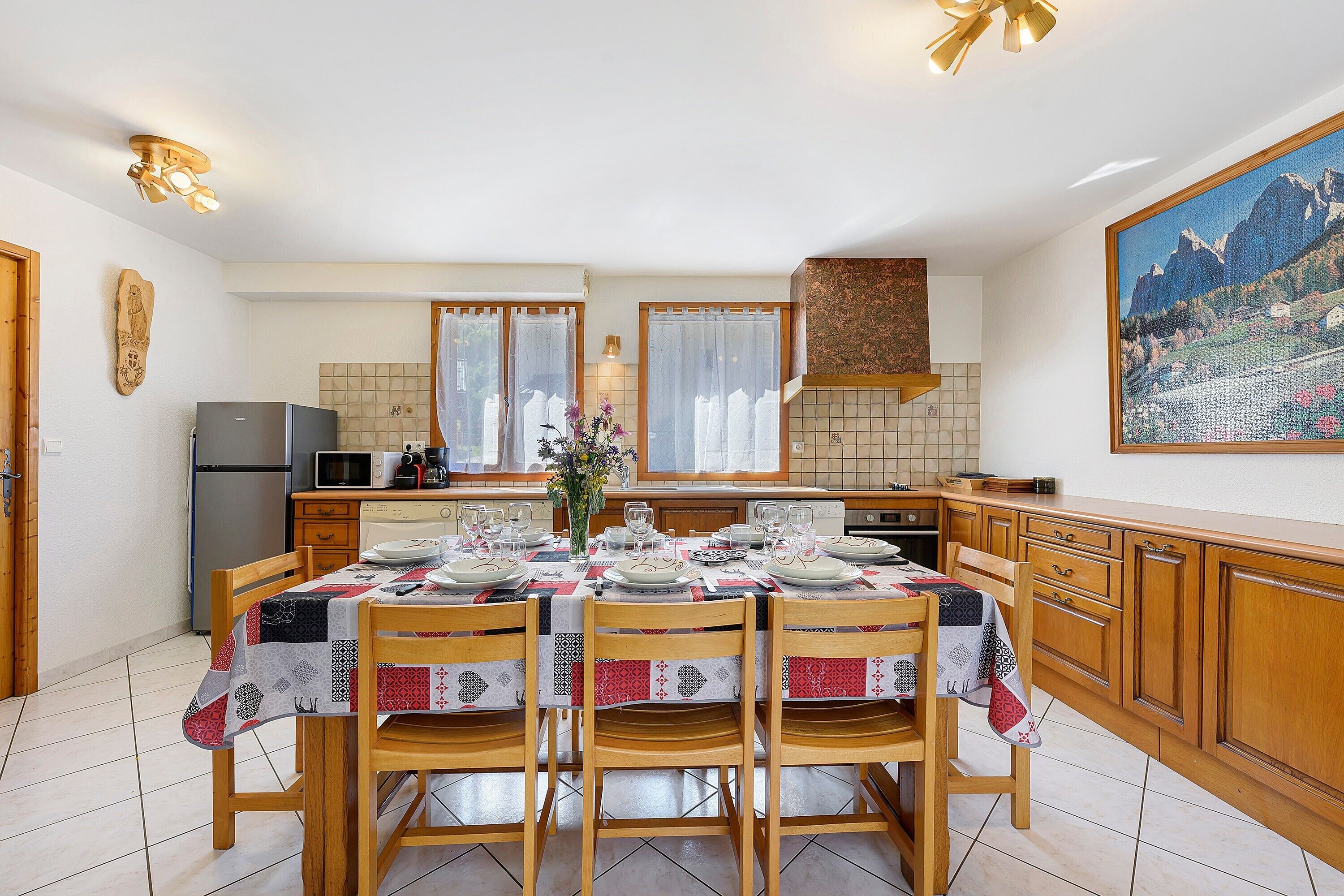Un coin repas avec une table dressée pour quatre personnes, des chaises en bois, une cuisine en arrière-plan et un tableau accroché au mur.