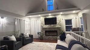 A spacious living room with a stone fireplace, a large TV, and a high ceiling with skylights.