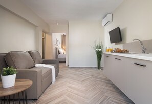 A modern living room with a sofa, a small table with a plant, and a kitchen area with a TV, sink, and breakfast items.