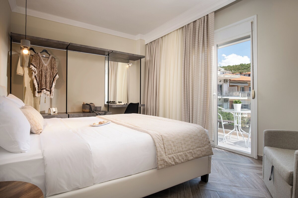 A hotel room with a bed, a desk, a chair, a balcony with a table and chairs, and a view of buildings.