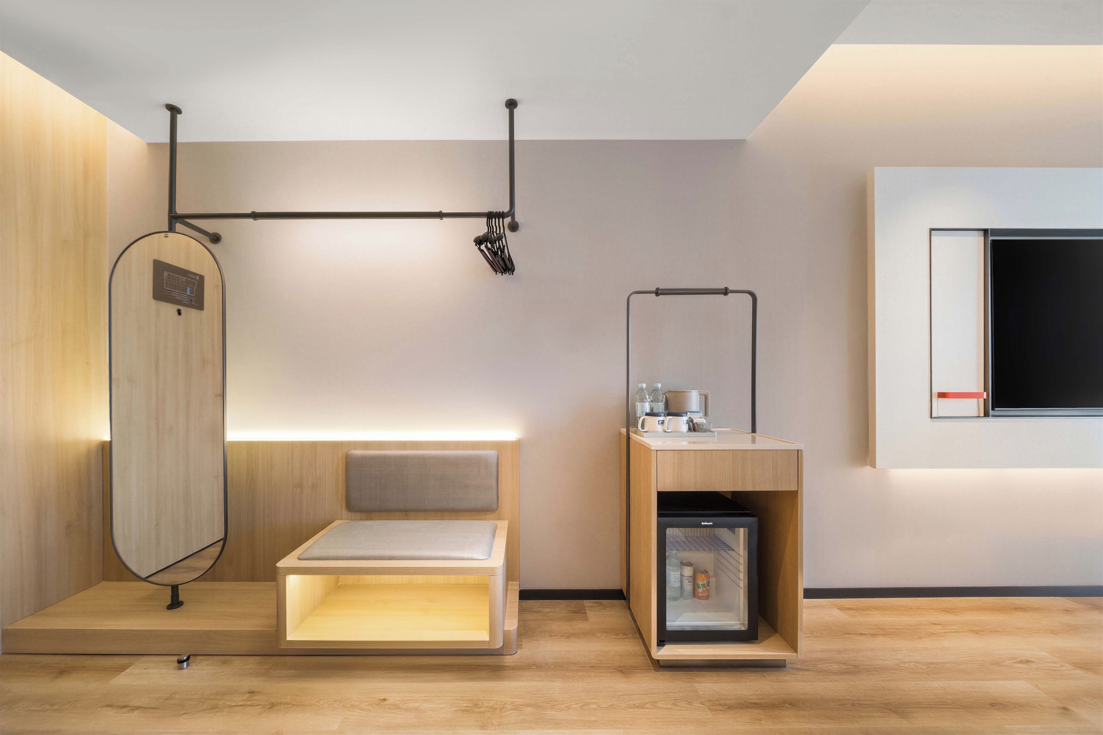 A modern hotel room with a wooden floor, a bed with a headboard, a small seating area, a minibar, and a TV mounted on the wall.