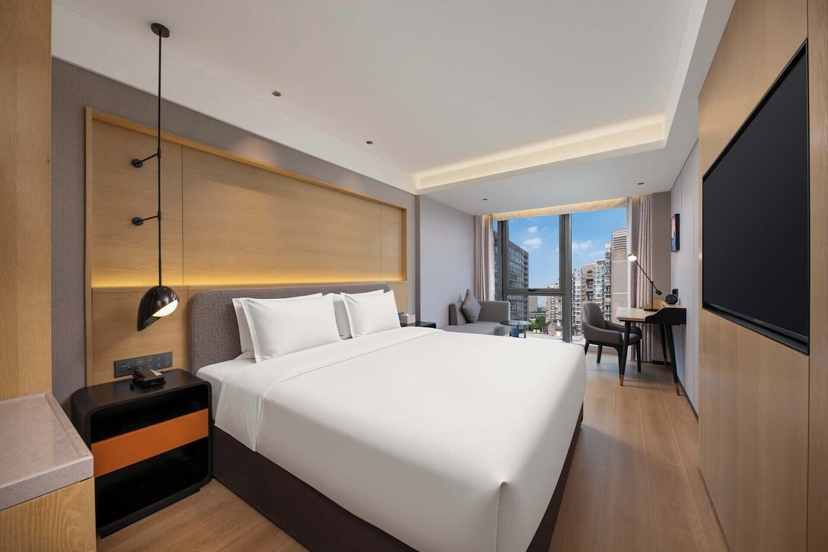 Habitación de hotel con una cama grande, mesita de noche, lámpara, escritorio, silla y un amplio ventanal con vistas a la ciudad.