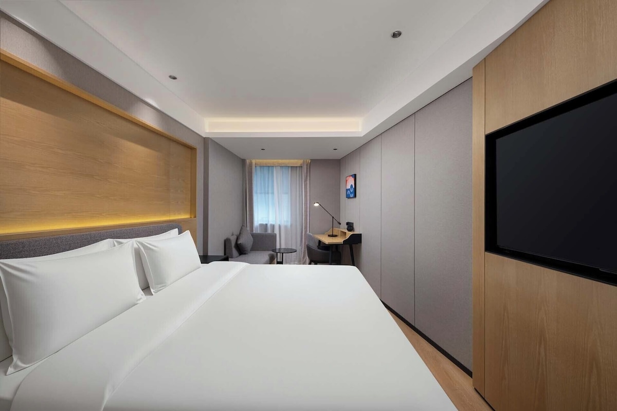 Una habitación de hotel con cama, escritorio, silla, televisor y ventana con cortinas.