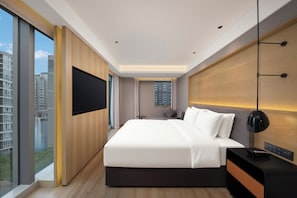 Quarto de hotel moderno com uma cama grande, uma televisão, uma mesa de cabeceira e vista para a cidade.