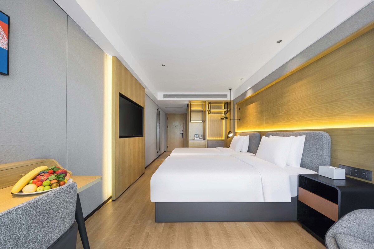 A modern hotel room with a large bed, a desk with fruit, a TV, and a bedside table with a lamp.