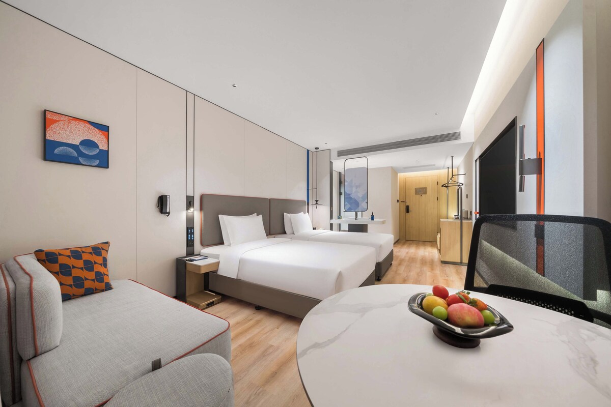 A hotel room with a bed, a desk, a chair, a small table with fruit, and a painting on the wall.