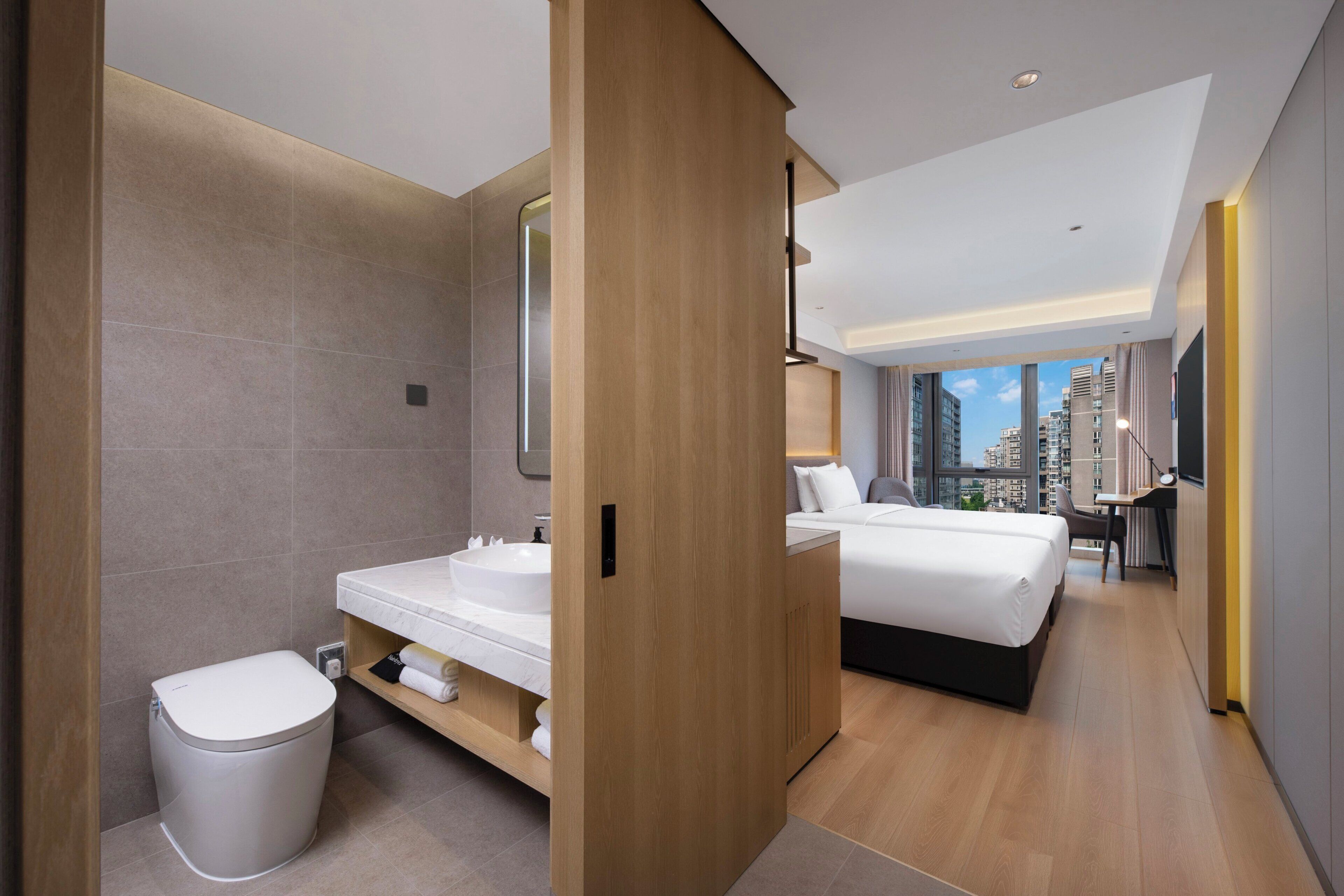 Habitación de hotel moderna con una cama grande, un baño con inodoro y lavamanos, y vista a la ciudad.