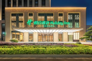 A modern hotel facade with illuminated signage, large windows, and a well-maintained garden area.