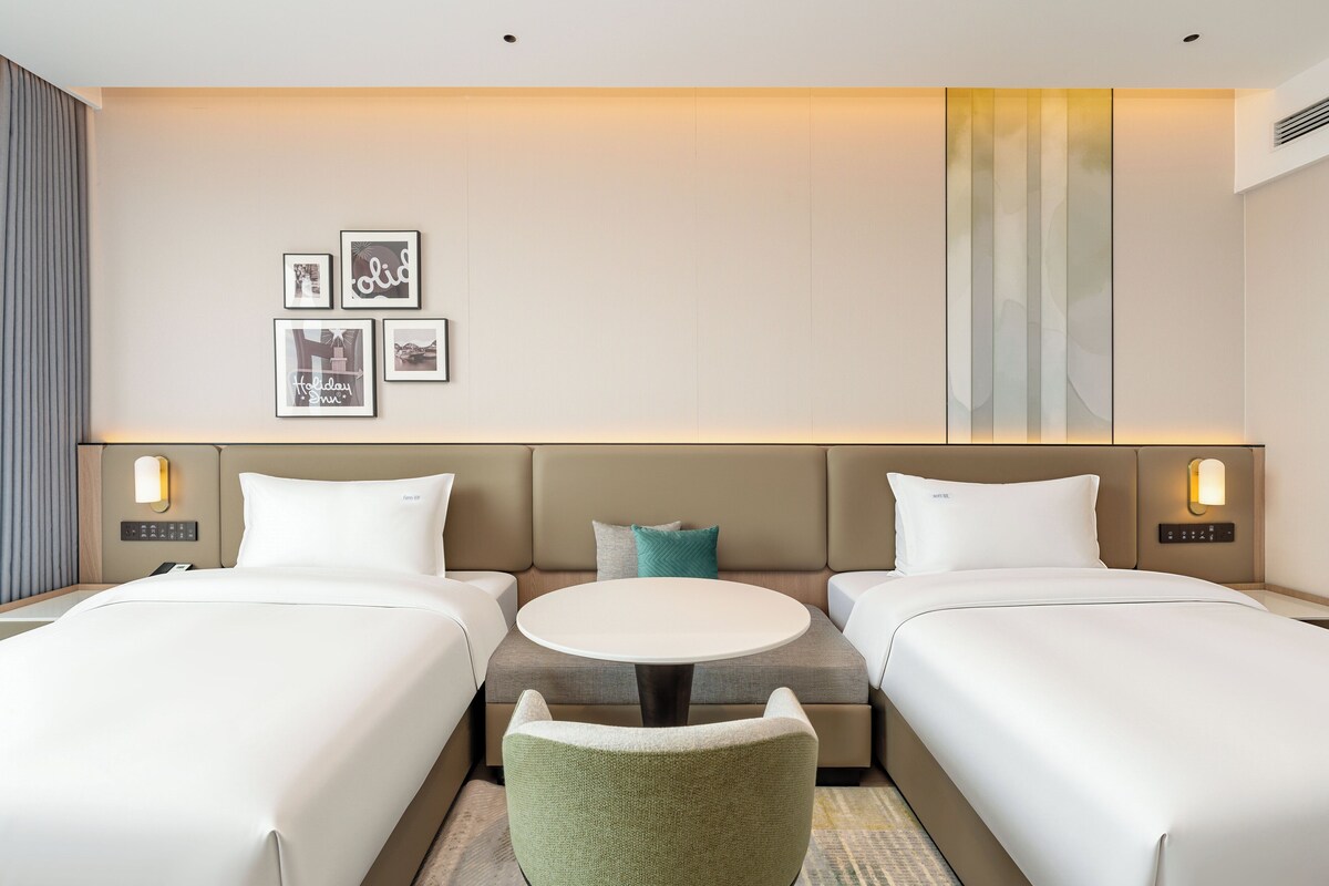 A hotel room with two beds, a sofa, a small table, and wall-mounted lights.
