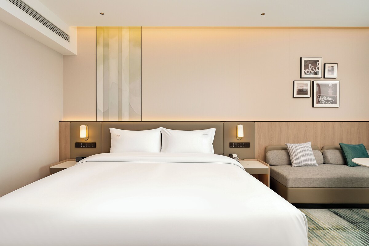 A hotel room with a large bed, bedside tables, a sofa, and wall-mounted lighting.