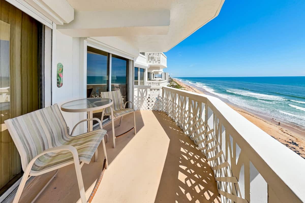 A balcony with a view of the ocean, a table, chairs, and a window with a curtain.