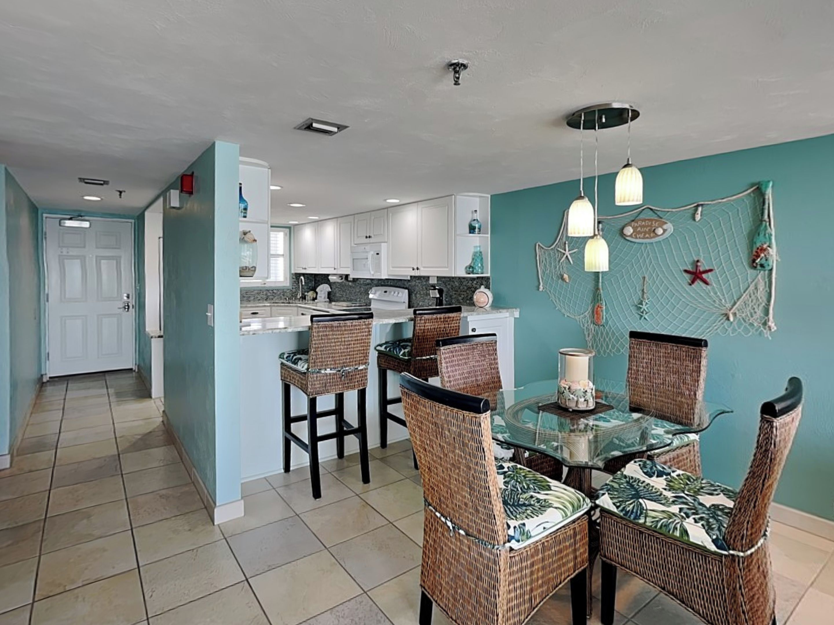 Una cocina con armarios blancos, un comedor con sillas de mimbre y una mesa de vidrio, y una pared verde azulado con una decoración de estrella de mar.