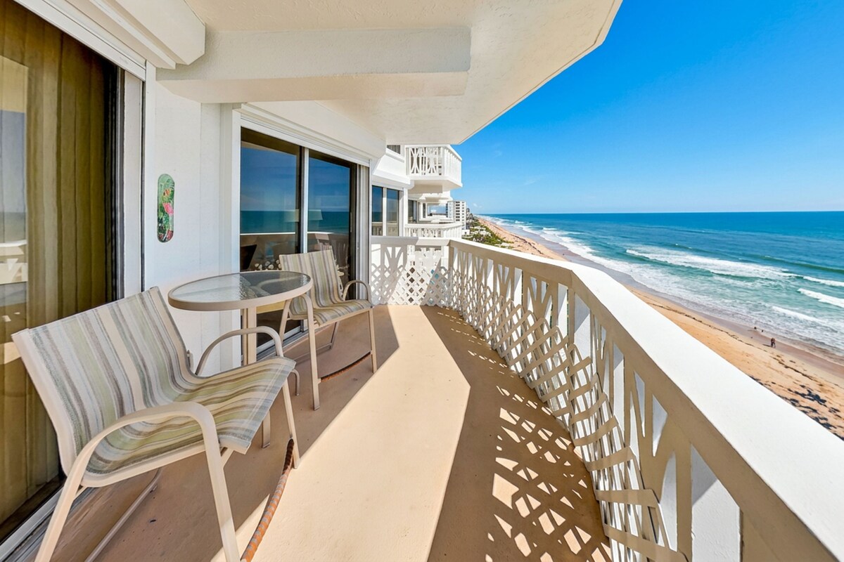 A balcony with a table and chairs, a view of the ocean, and a building with a sign.