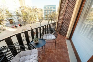 Ein Balkon mit zwei gepolsterten Stühlen, einem kleinen Tisch mit einer Pflanze und Blick auf eine Straße mit Gebäuden und Bäumen.