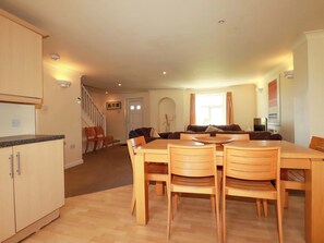 A dining area with a wooden table and chairs, a kitchen with cabinets, a staircase, and a living room with a sofa and a window. - 2 Atlantic Reach (St. Columb Major)