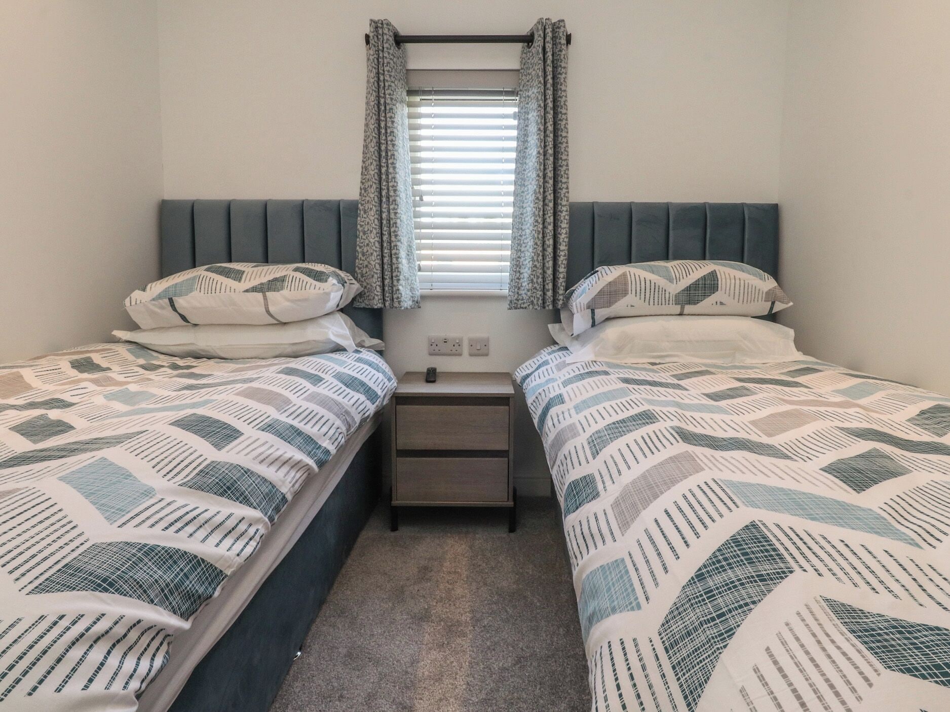 A twin room with two beds, a window with blinds, a bedside table, and a power outlet.