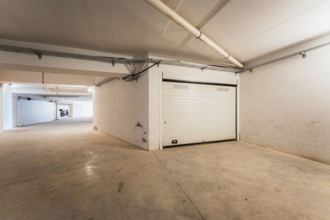 A large, empty parking garage with concrete floors and walls, a closed garage door, and visible pipes on the ceiling.