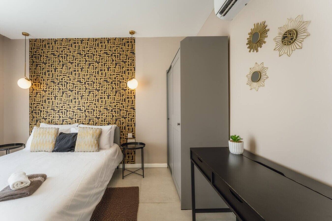Une chambre d’hôtel moderne avec un papier peint à motifs, un lit avec des oreillers, une table de chevet, un bureau, une petite plante et des décorations murales.