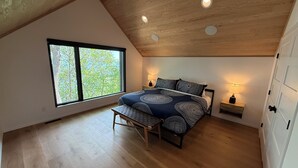 Un dormitorio con un ventanal grande, una cama con ropa de cama estampada, lámparas de noche, un banco y techo de madera.