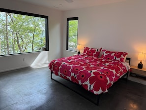 Un dormitorio con una cama grande, dos lámparas de noche, una ventana con vistas a árboles y un suelo de hormigón.