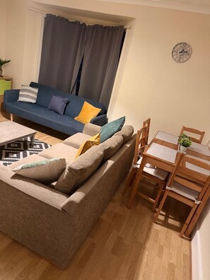 Ein Wohnzimmer mit einer Couch, einem Esstisch mit Stühlen, einer Wanduhr und einem Fenster mit Vorhängen.