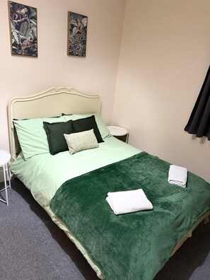 Ein Bett mit grüner Decke, Kissen und einem weißen Kopfteil. Darüber an der Wand hängen zwei Gemälde.