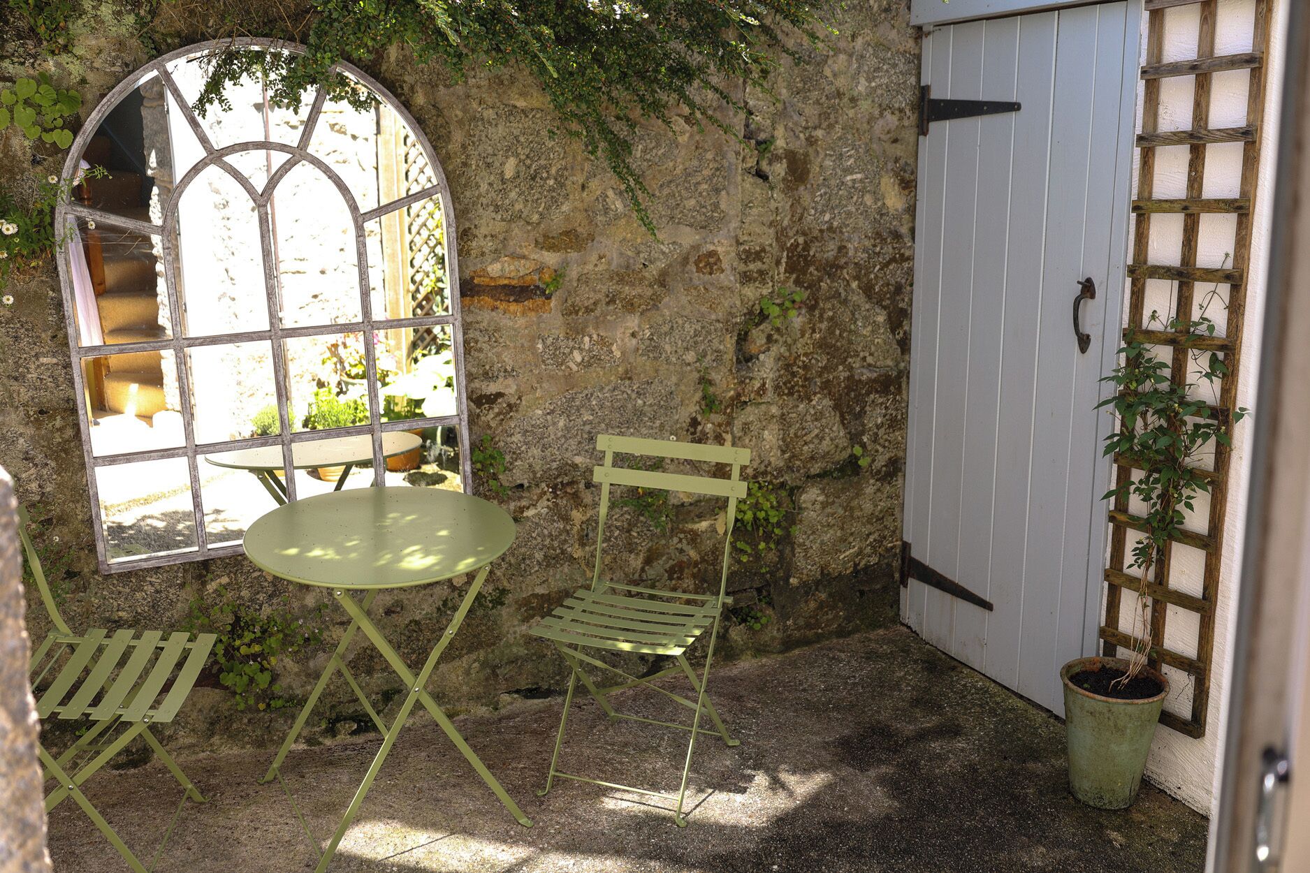 Un muro de piedra rústico, una mesa y una silla de metal verde, una ventana con vistas y una puerta blanca con celosía.