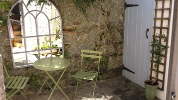 Un muro de piedra rústico, una mesa y una silla de metal verde, una ventana con vistas y una puerta blanca con celosía.