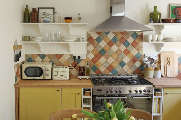A kitchen with a stove, microwave, and vintage toaster. There are shelves above holding dishes and bottles. - Kittiwake (Penzance)