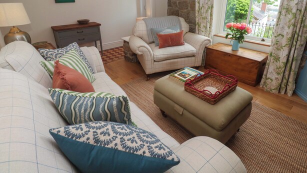 Ein Wohnzimmer mit Sofa, Sessel, Hocker und einem kleinen Tisch mit einem Buch und einem Korb.
